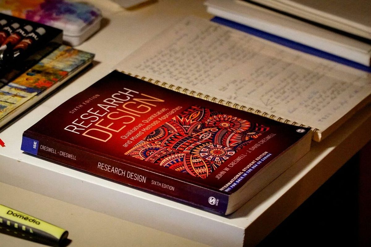 Creswell's Research Design lying on a desk beside an open notebook of handwritten notes, a highlighter and a paint box.