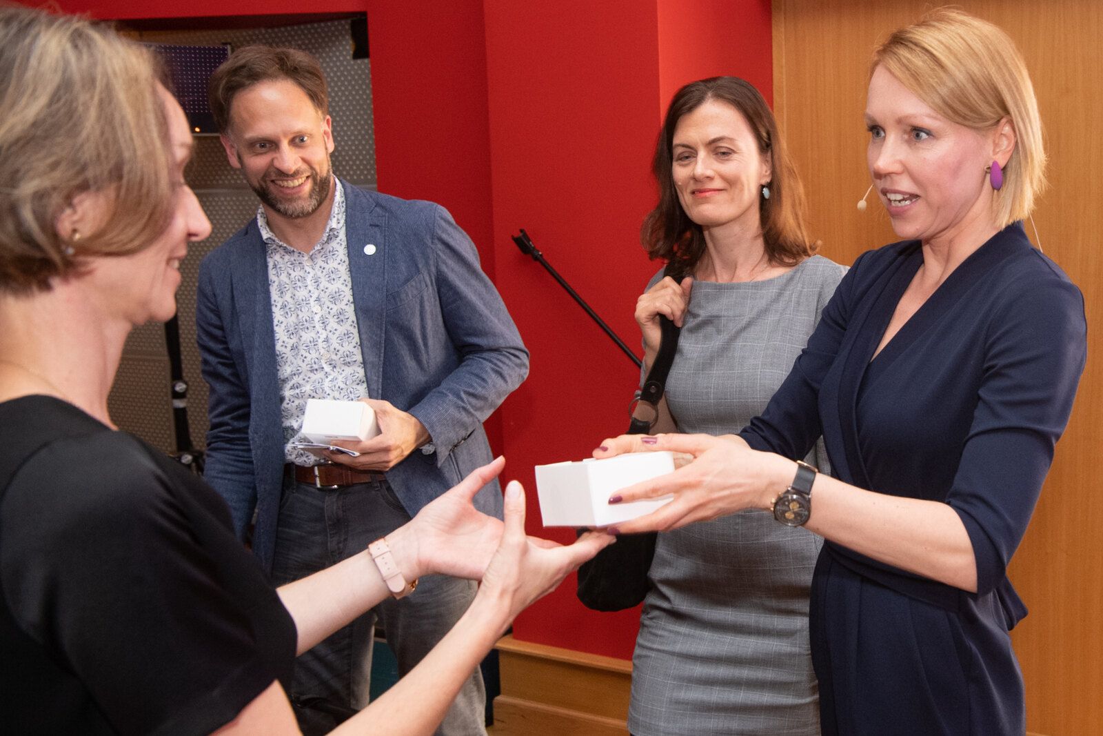 After an event: four people talking, two of them handing over small white boxes, against a red wall.