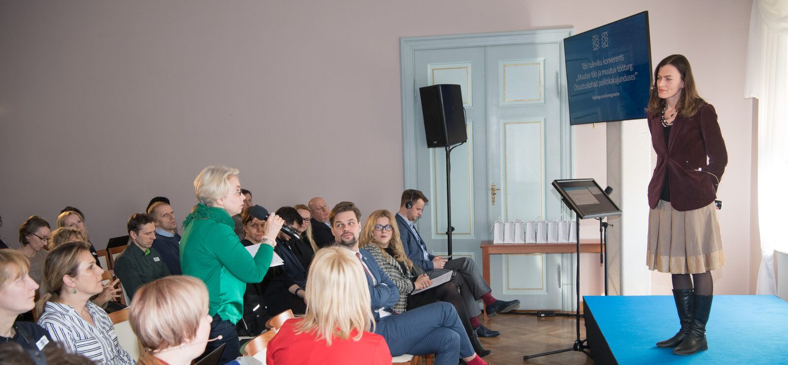 A conference room in daylight: a seated audience at the left, a woman standing among them with a microphone to ask a question, and a speaker on a low blue stage at the right, listening.